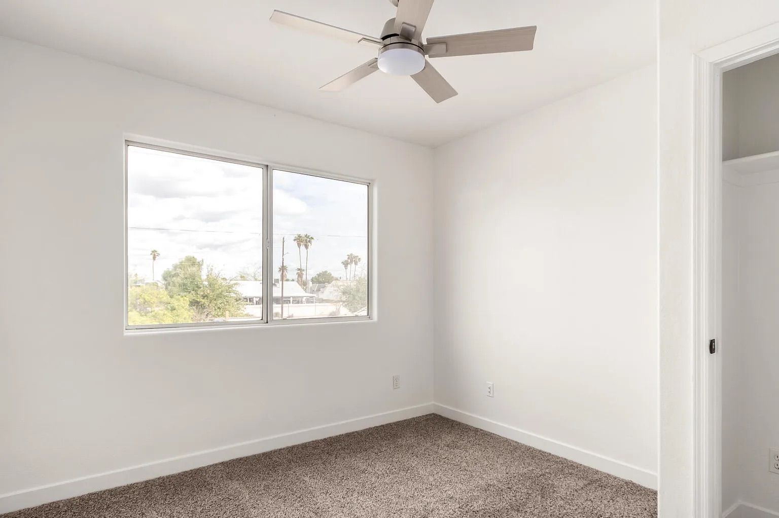 Empty bedroom space with tan carpeting, white walls, a ceiling fan, a view from the window, and an open closet space.