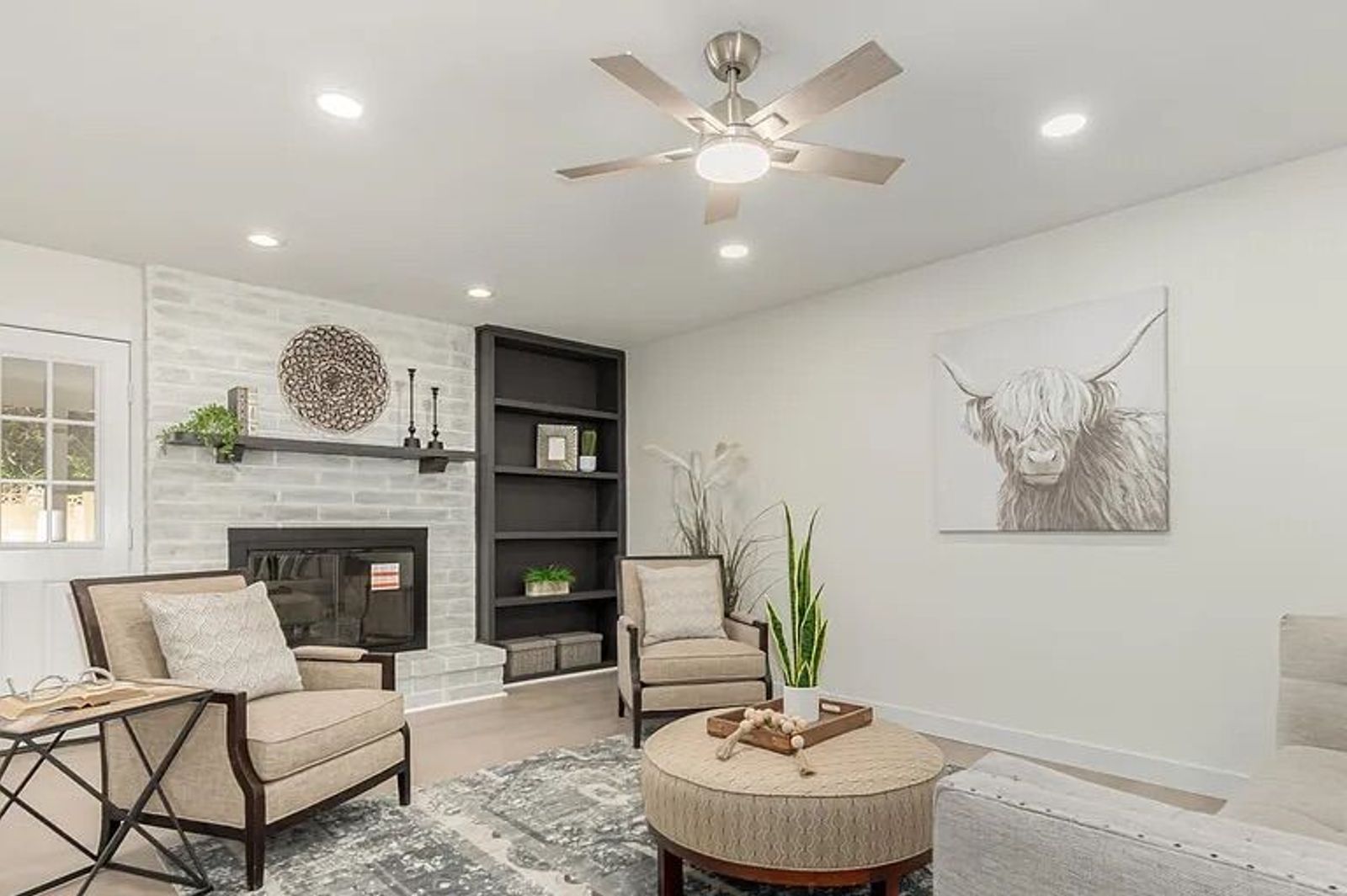 Cozy living room showing a whitewashed brick fireplace with a black mantel, dark built-in bookshelves, beige armchairs, and a round tufted ottoman.