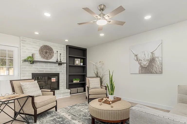 Cozy living room showing a whitewashed brick fireplace with a black mantel, dark built-in bookshelves, beige armchairs, and a round tufted ottoman.