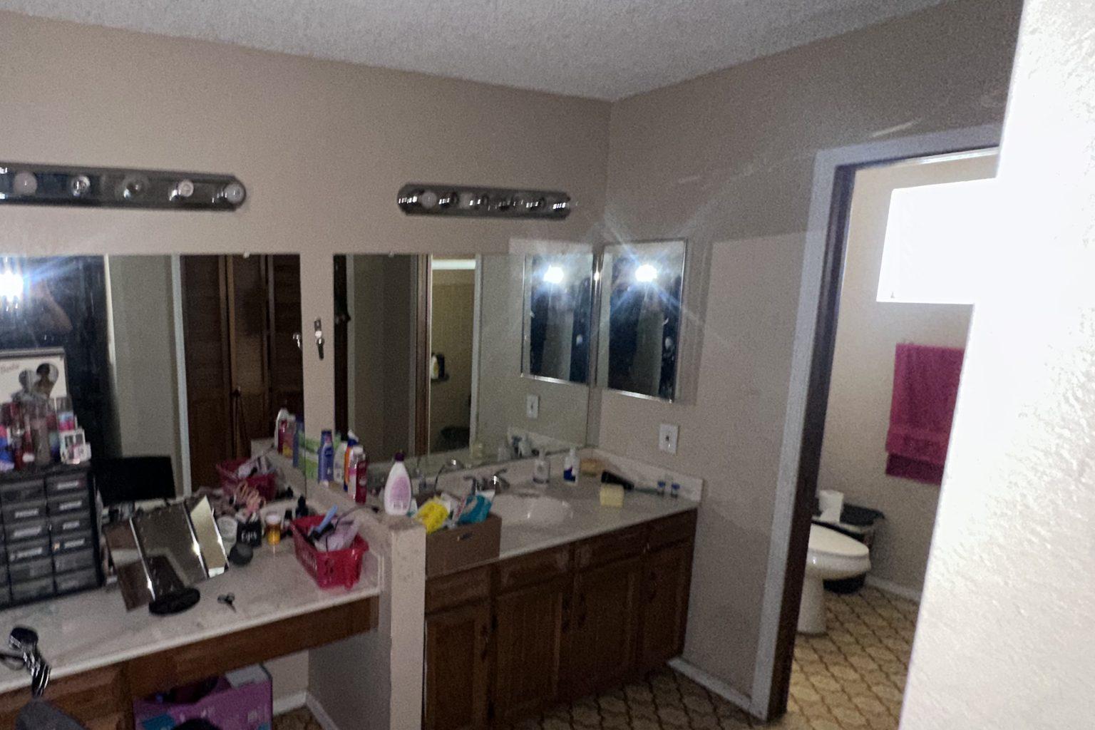 Before the remodel: the original primary bathroom with a long wood vanity, strip lighting, wide mirrors and the water closet beyond.