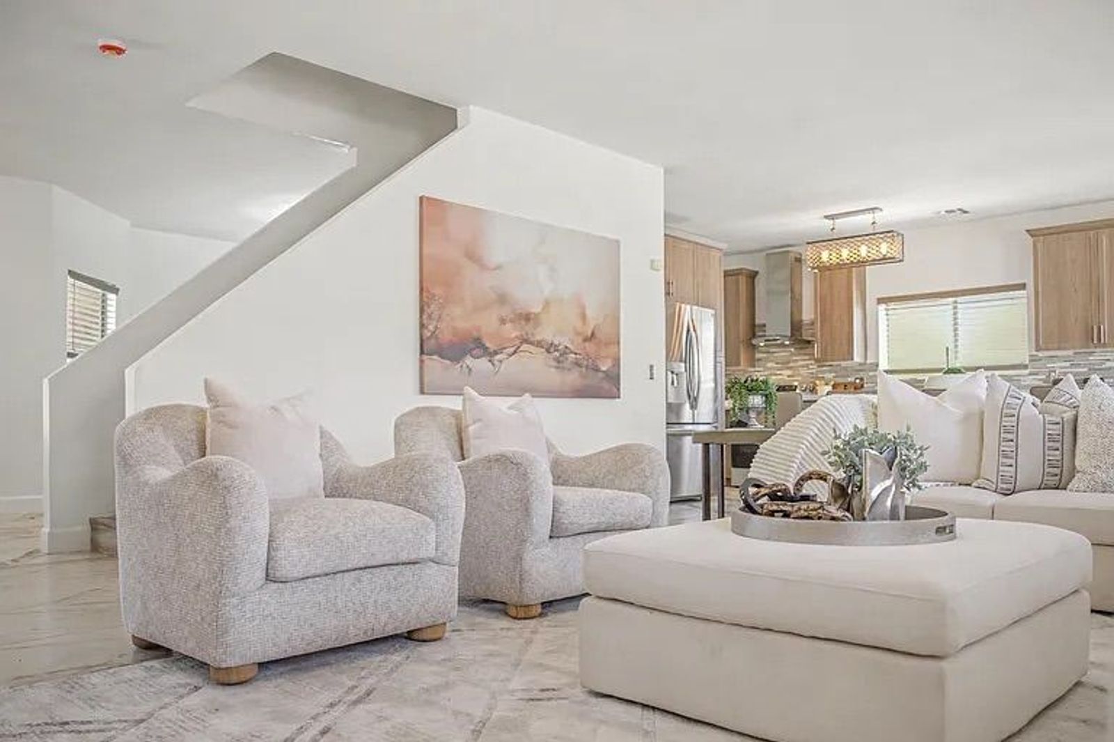 View from the living area showing two textured armchairs, a white ottoman, marble-look flooring, and a white staircase leading to the second floor.