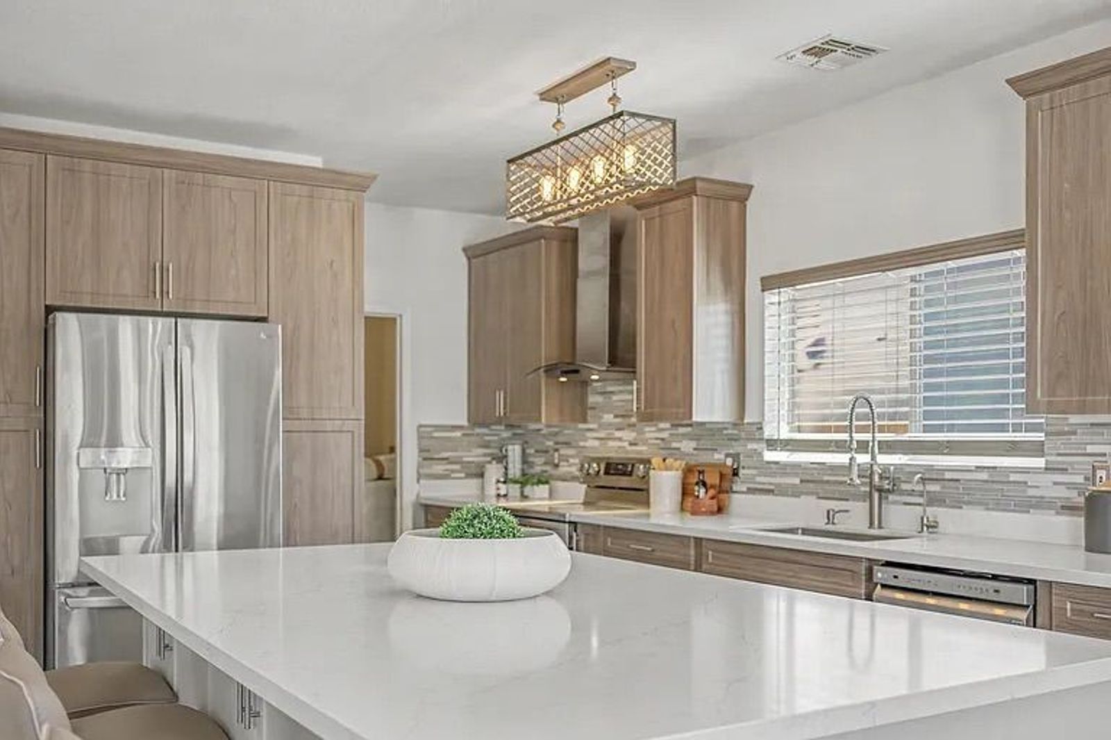 Modern kitchen featuring a large white quartz island with beige barstools, light wood cabinetry, stainless steel appliances, and a unique rectangular wire chandelier.