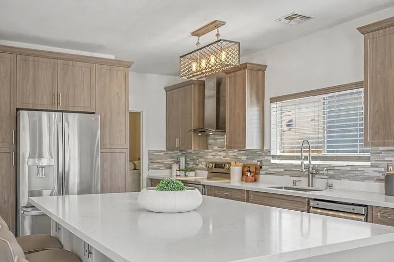 Modern kitchen featuring a large white quartz island with beige barstools, light wood cabinetry, stainless steel appliances, and a unique rectangular wire chandelier.