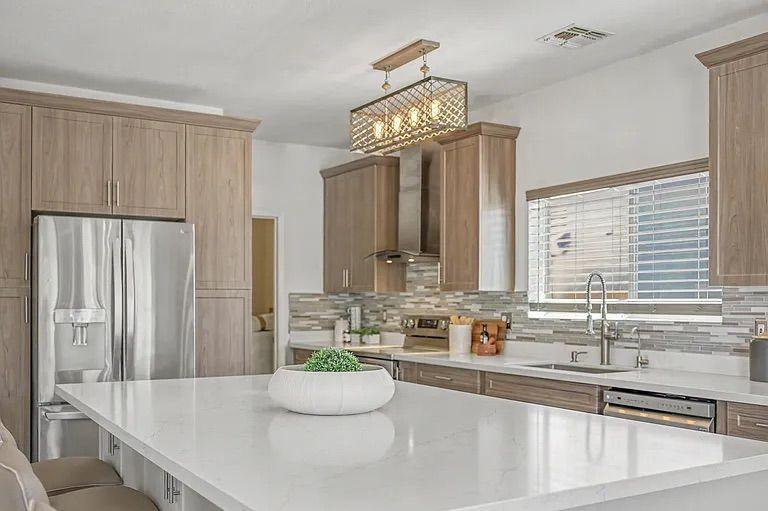 Modern kitchen featuring a large white quartz island with beige barstools, light wood cabinetry, stainless steel appliances, and a unique rectangular wire chandelier.
