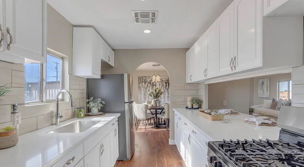 Bright kitchen featuring white cabinetry, light countertops, stainless steel appliances, dark wood flooring, and an arched doorway leading to a formal dining room.
