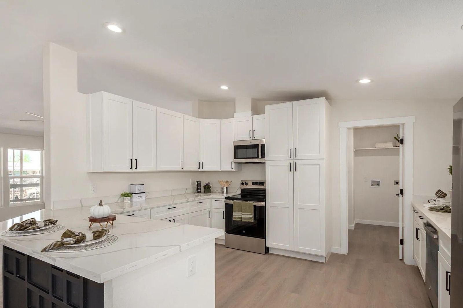 Modern kitchen displaying white shaker cabinets, quartz countertops, a black geometric island, stainless steel appliances, and access to a laundry room.