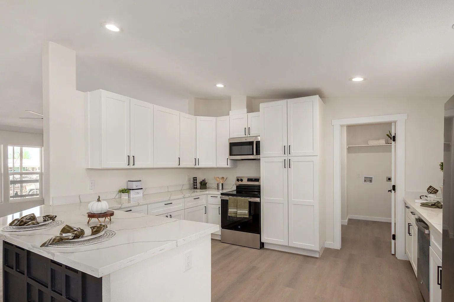 Modern kitchen displaying white shaker cabinets, quartz countertops, a black geometric island, stainless steel appliances, and access to a laundry room.