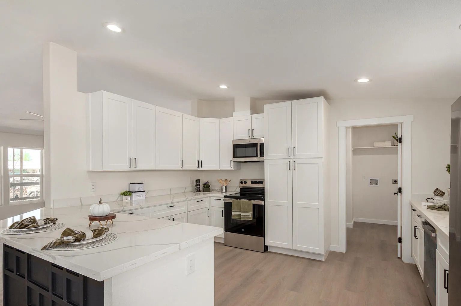 Modern kitchen displaying white shaker cabinets, quartz countertops, a black geometric island, stainless steel appliances, and access to a laundry room.