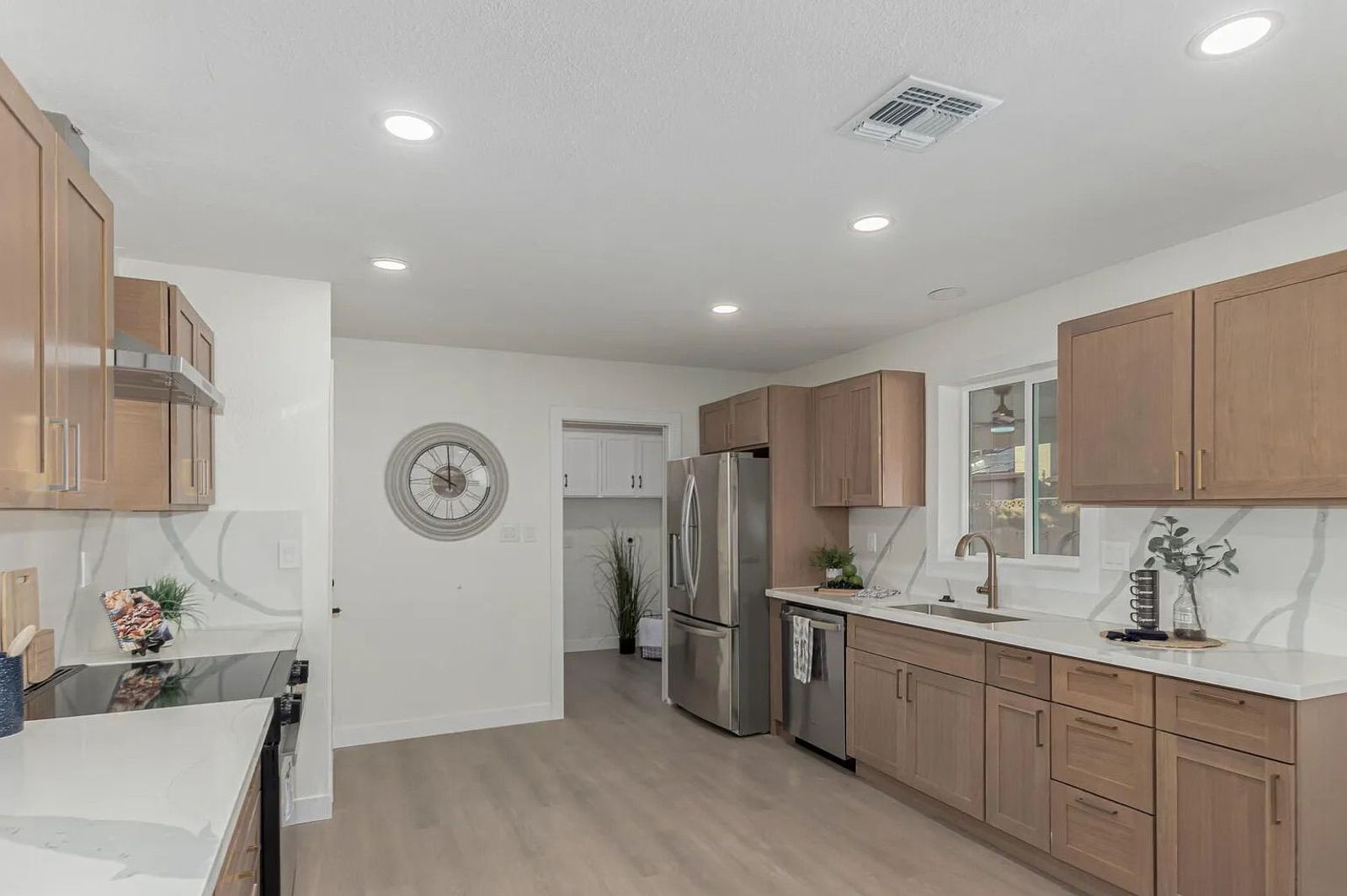 Modern kitchen featuring light wood shaker cabinets, white quartz countertops, stainless steel appliances, light wood-look flooring, and a large decorative wall clock.