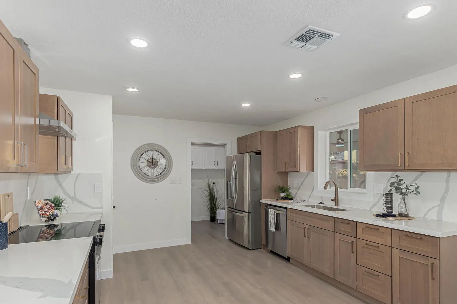 Modern kitchen featuring light wood shaker cabinets, white quartz countertops, stainless steel appliances, light wood-look flooring, and a large decorative wall clock.