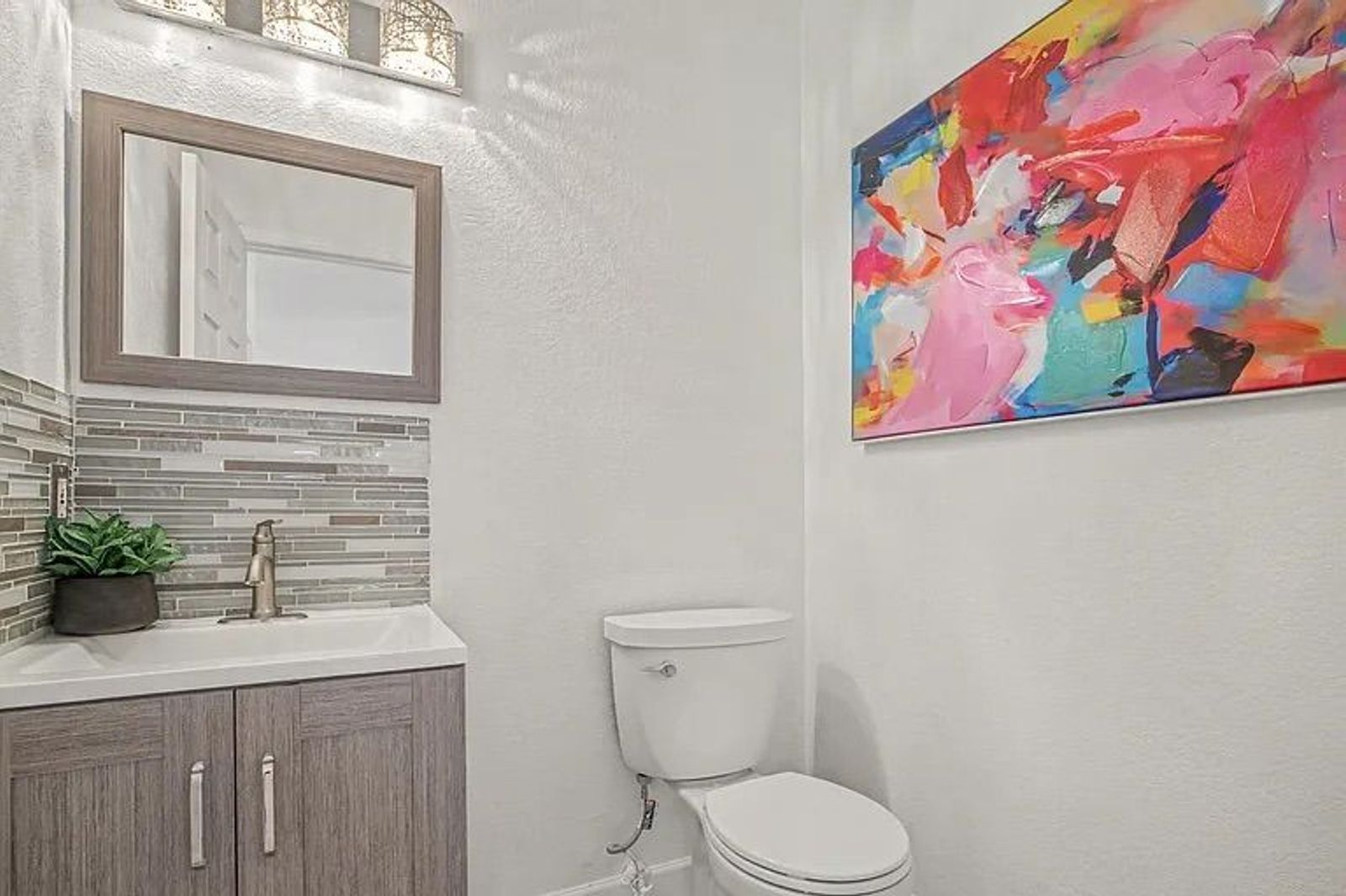 Small powder room featuring a grey wood-look vanity, a glass tile backsplash, a white toilet, and a vibrant abstract canvas painting on the wall.