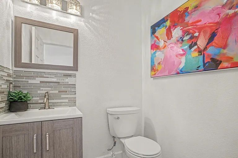 Small powder room featuring a grey wood-look vanity, a glass tile backsplash, a white toilet, and a vibrant abstract canvas painting on the wall.