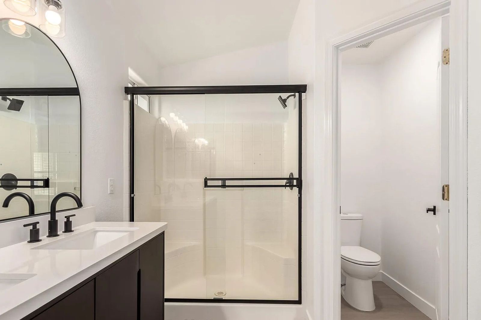 View of a bathroom featuring a dark double vanity, a glass-enclosed step-in shower with white tile, and an adjacent open water closet.