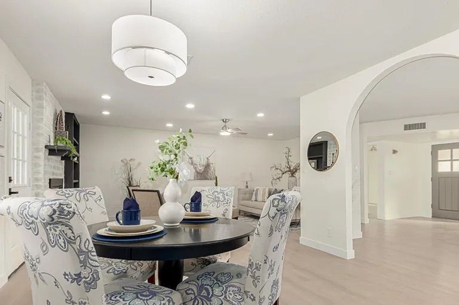Dining space featuring a round black table with blue floral patterned chairs, a modern drum chandelier, and a view into a living room with a whitewashed brick fireplace.