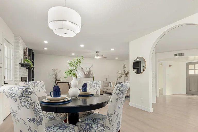 Dining space featuring a round black table with blue floral patterned chairs, a modern drum chandelier, and a view into a living room with a whitewashed brick fireplace.