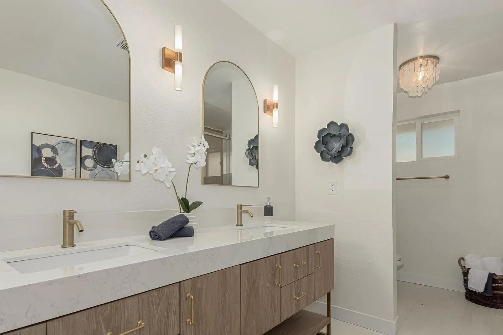 Bathroom vanity area featuring a textured wood double vanity, white quartz countertops, gold hardware, two arched mirrors with gold wall sconces, and a partial view of a water closet.