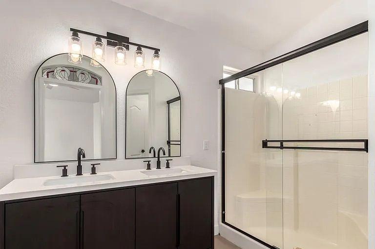 Bathroom showing a dark wood double vanity with white countertops, dual arched mirrors, black hardware, and a glass-enclosed white tile shower.