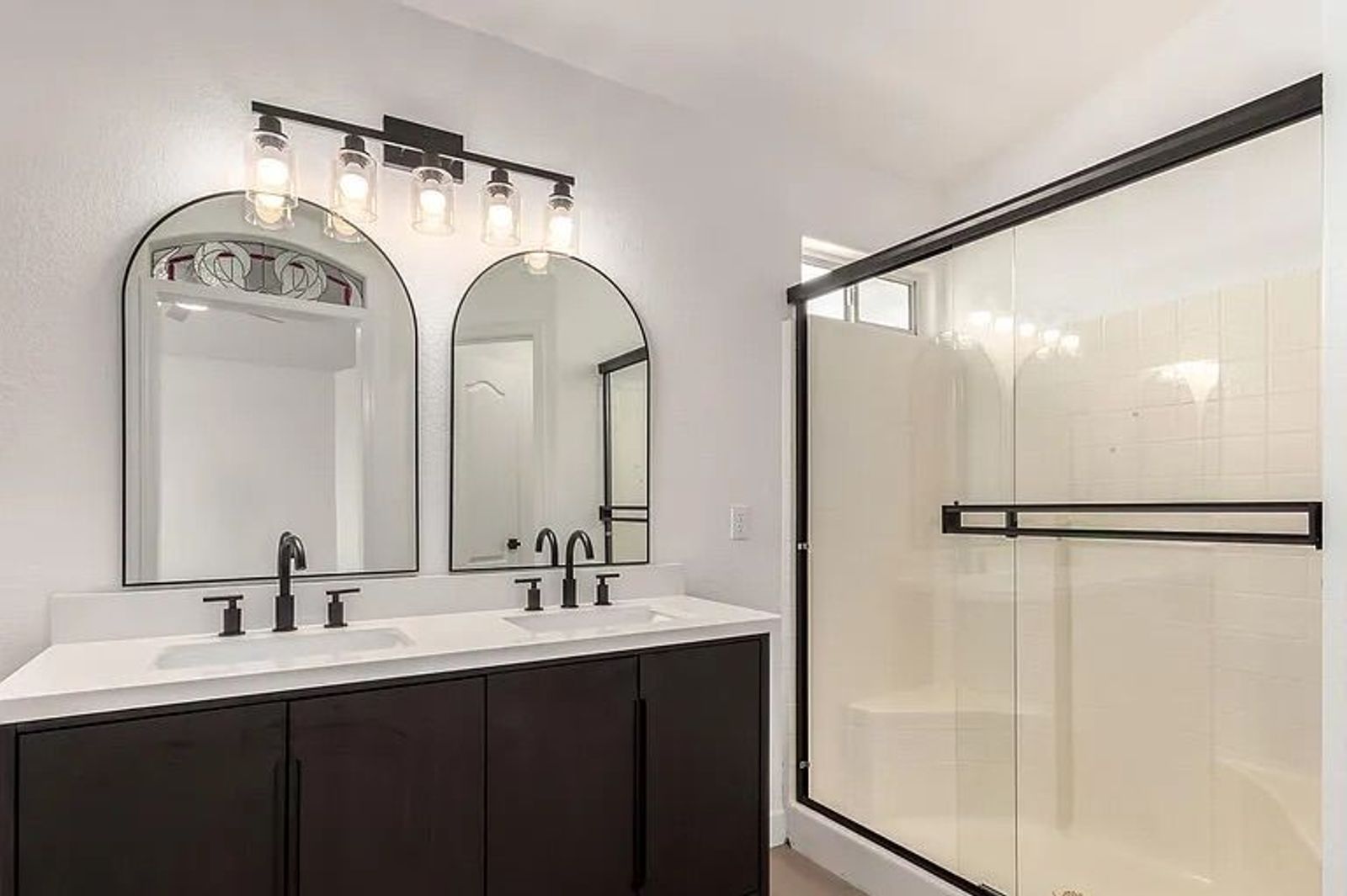 Bathroom showing a dark wood double vanity with white countertops, dual arched mirrors, black hardware, and a glass-enclosed white tile shower.