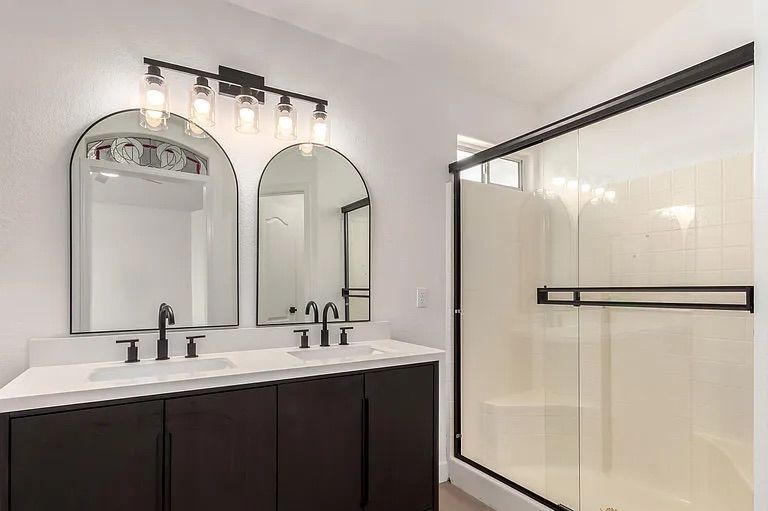 Bathroom showing a dark wood double vanity with white countertops, dual arched mirrors, black hardware, and a glass-enclosed white tile shower.