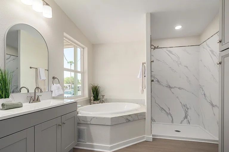 Primary bathroom highlighting a grey dual-sink vanity, arched mirrors, a white corner soaking tub, and a walk-in shower with marble-look panels.