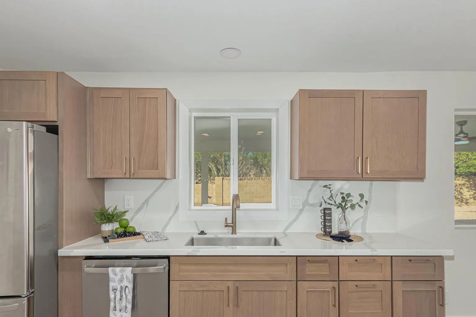 Kitchen interior showing light wood shaker cabinets, a stainless steel dishwasher, and white quartz countertops that seamlessly extend up the wall as a backsplash.