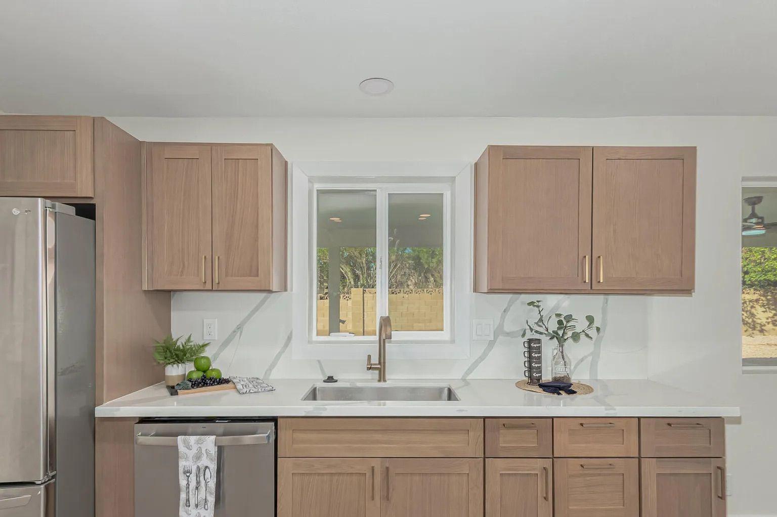 Kitchen interior showing light wood shaker cabinets, a stainless steel dishwasher, and white quartz countertops that seamlessly extend up the wall as a backsplash.