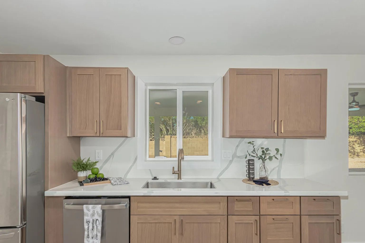 Kitchen interior showing light wood shaker cabinets, a stainless steel dishwasher, and white quartz countertops that seamlessly extend up the wall as a backsplash.
