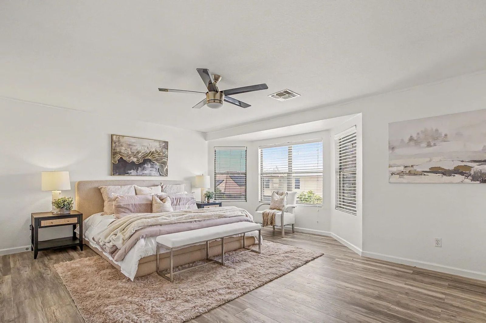 Spacious primary bedroom featuring light wood-style flooring, a large bed with blush and white bedding, a bay window seating area, and a textured area rug.