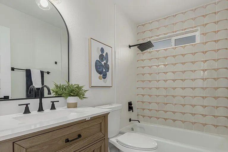 Bathroom highlighting a wood vanity with a black faucet, a large arched mirror, and a bathtub/shower combination featuring a pink and white scallop-patterned tile surround and matte black fixtures.