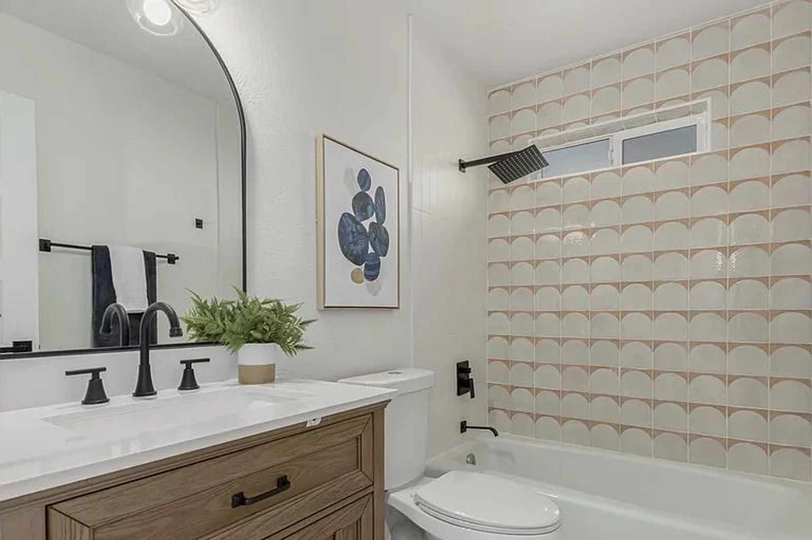 Bathroom highlighting a wood vanity with a black faucet, a large arched mirror, and a bathtub/shower combination featuring a pink and white scallop-patterned tile surround and matte black fixtures.