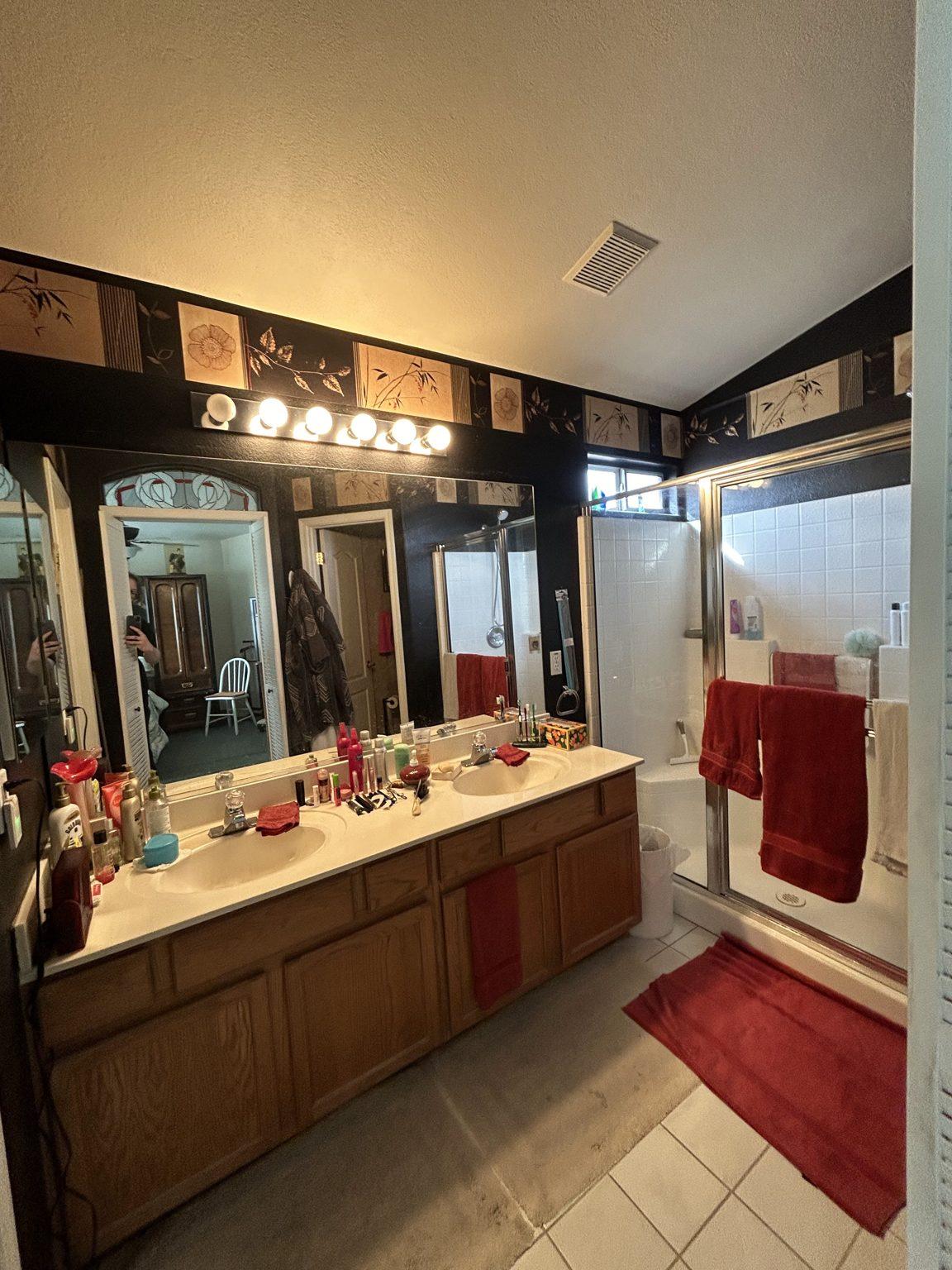 Before the remodel: the original primary bathroom with an oak double vanity, strip lighting, dark wallpaper and a shower at right.