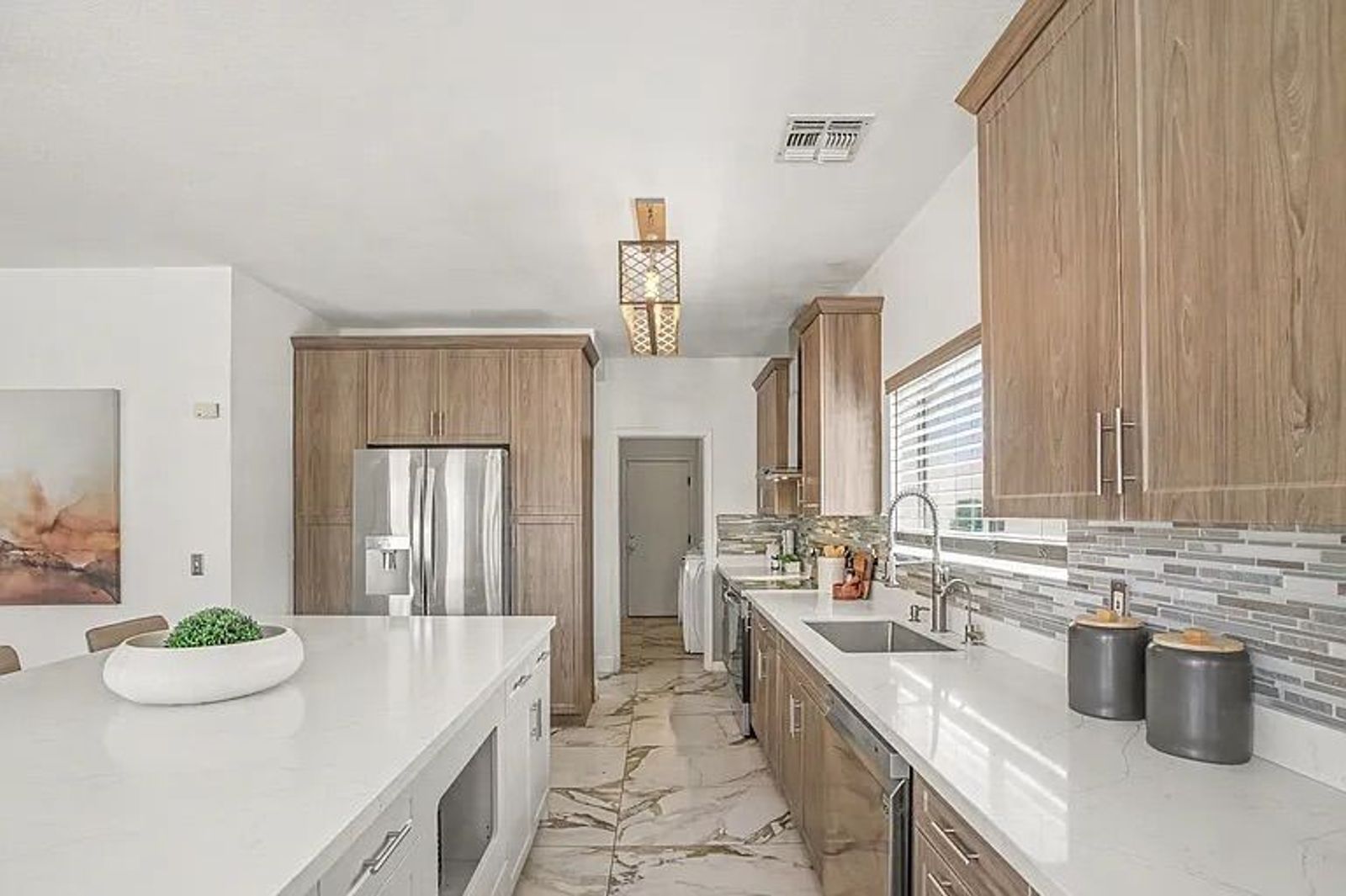 View down a bright kitchen showcasing light wood-finish cabinetry, a long white center island, marble-patterned tile flooring, and a doorway to a laundry room.