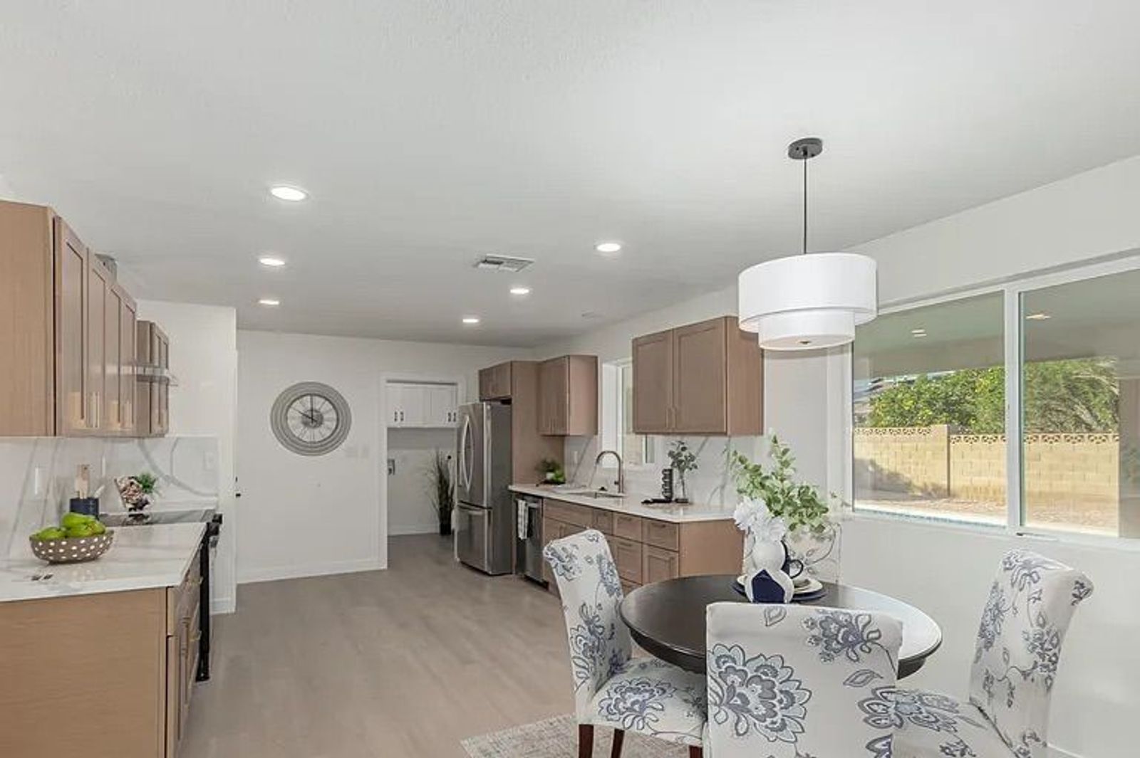 Kitchen and dining nook featuring light wood cabinetry, a round dining table with blue floral chairs, a modern white pendant light, and a large window looking out to the backyard pool.