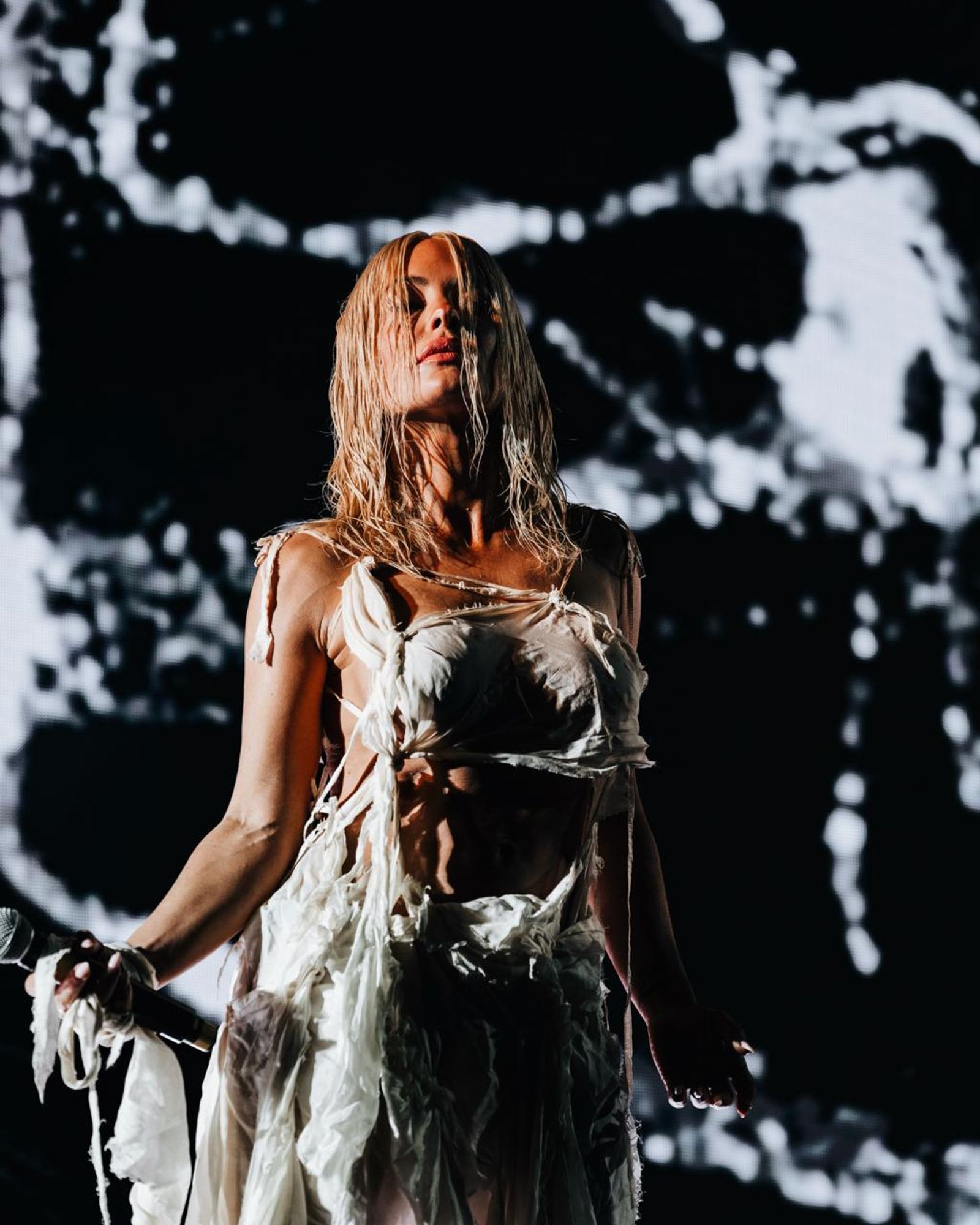 Aimée Sophia performing live, microphone in hand, against a projected monochrome cymatic field