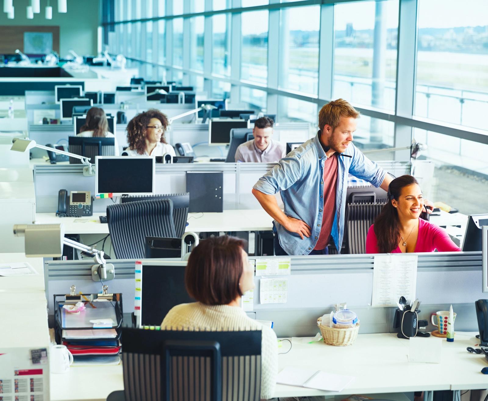 Busy open-plan office with staff at desks and two colleagues chatting between workstations
