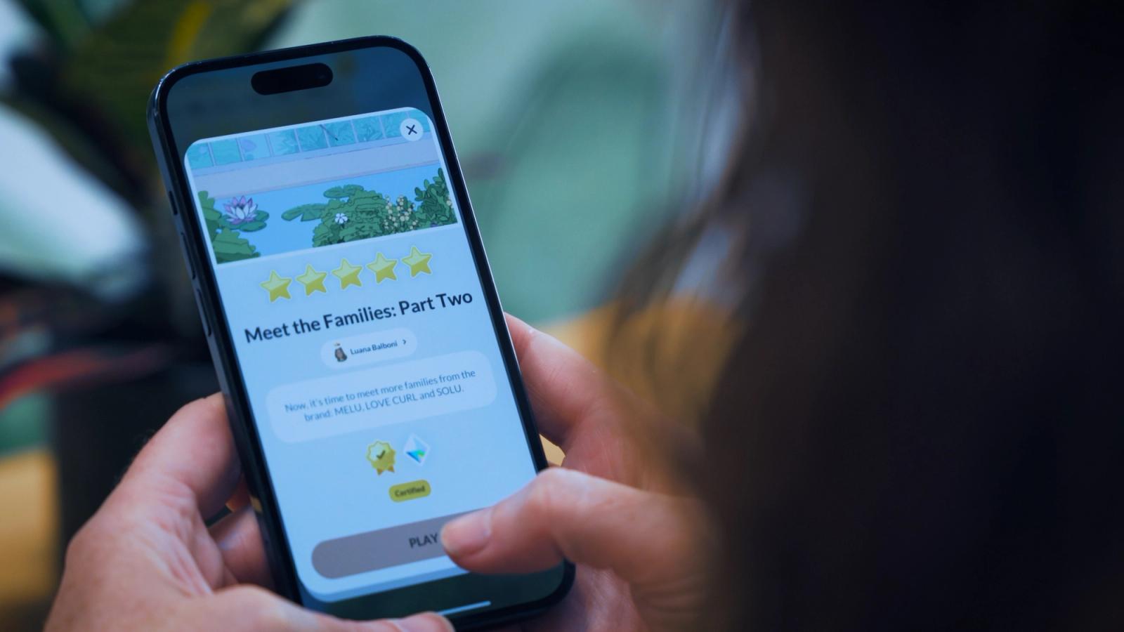 Hands hold a smartphone displaying an app titled "Meet the Families: Part Two" with five stars and a "Play" button.