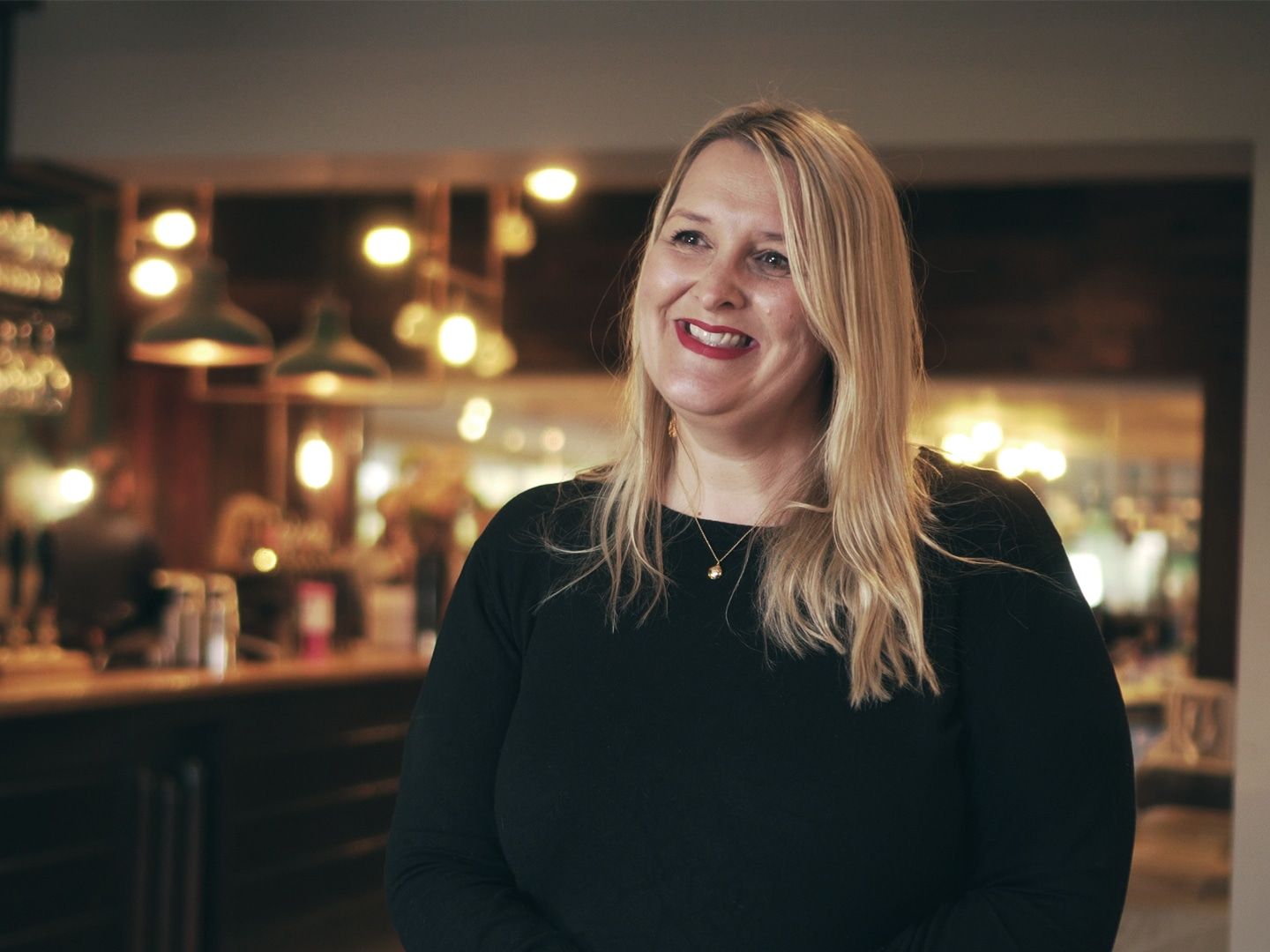 Jane Murray lächelt in schwarzem Oberteil in einem Pub, hinter ihr Hängeleuchten und die Bar