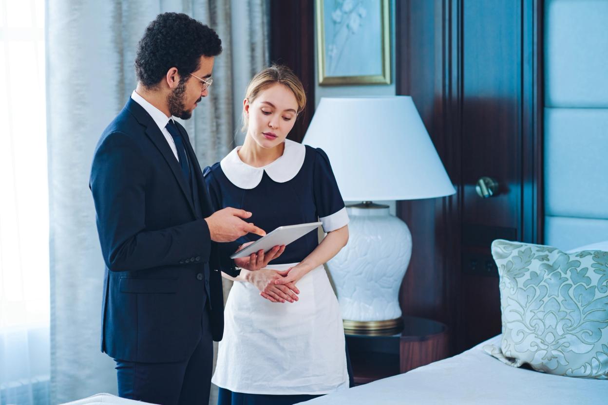 Selbstbewusster Besitzer eines Luxushotels gibt einer Housekeeping-Mitarbeiterin Anweisungen und zeigt auf einen Tablet-Bildschirm mit den Dienstpflichten des Servicepersonals
