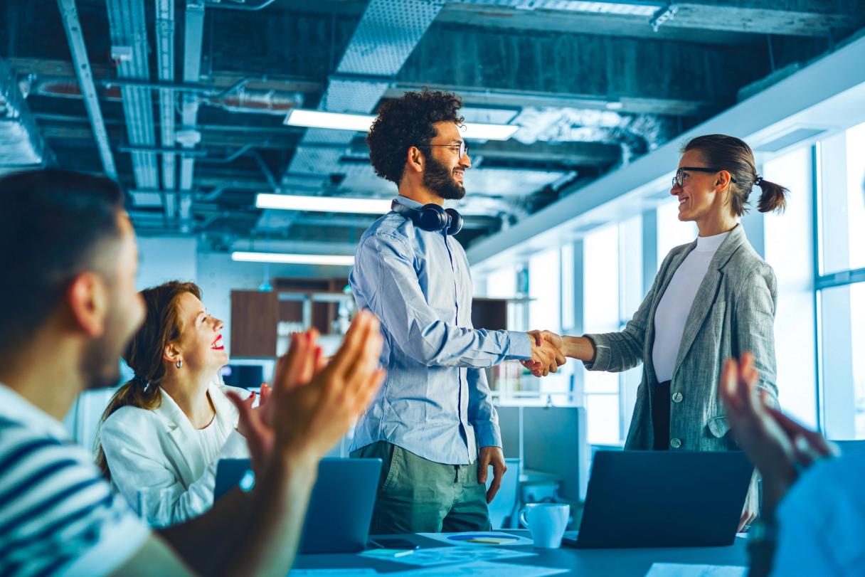 Zwei lächelnde Personen geben sich in einem Büro die Hand, während Kolleginnen und Kollegen applaudieren.