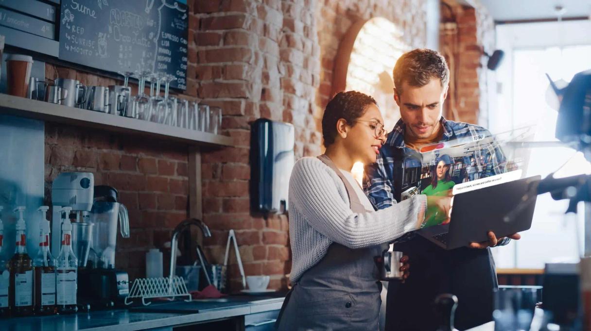 Deux employés de café regardent un ordinateur portable affichant une visioconférence holographique.