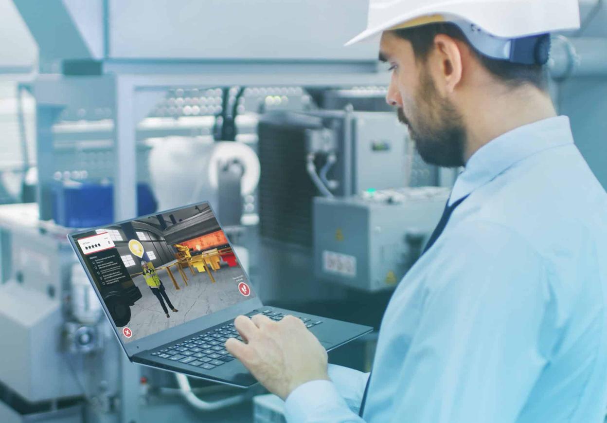 Arbeiter mit Schutzhelm betrachtet in einer Industrieanlage eine 3D-Sicherheitssimulation auf einem Laptop