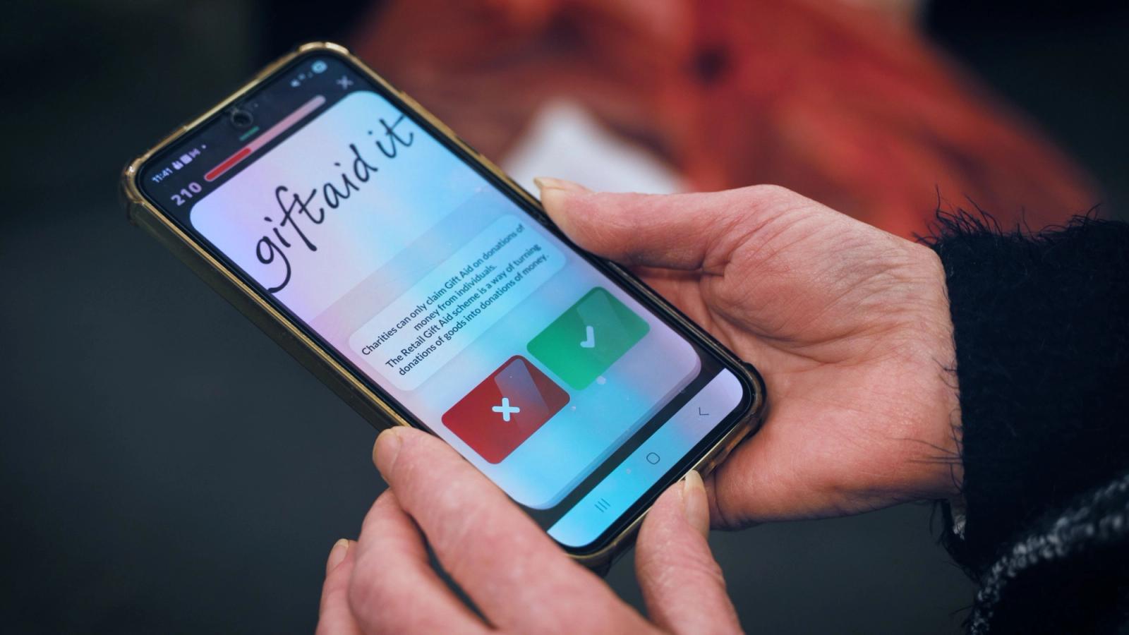 Hände halten ein Smartphone mit einem Quizbildschirm mit der Überschrift „giftaid it“ und roten Kreuz- und grünen Häkchen-Schaltflächen