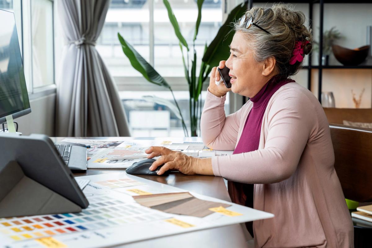 Ältere Mitarbeiterin telefoniert am Schreibtisch und arbeitet mit Farbmustern am Computer