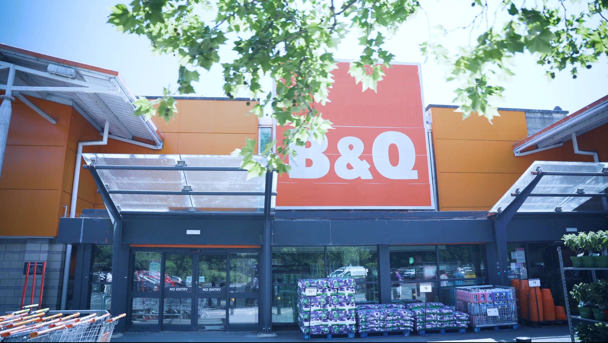 Außenansicht eines B&Q-Markts mit oranger Fassade und großem Logo über dem Eingang, umrahmt von Baumblättern.
