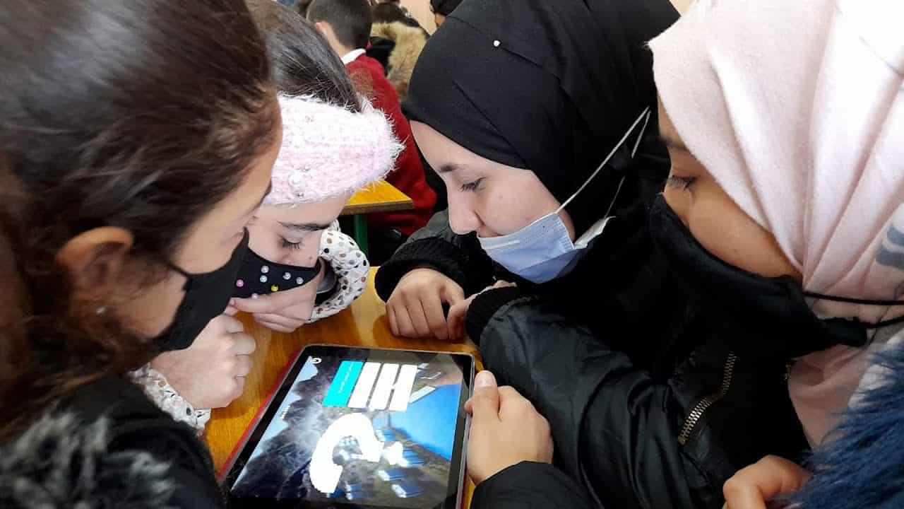 Mädchen mit Kopftüchern und Mund-Nasen-Masken versammeln sich im Klassenzimmer um ein Tablet