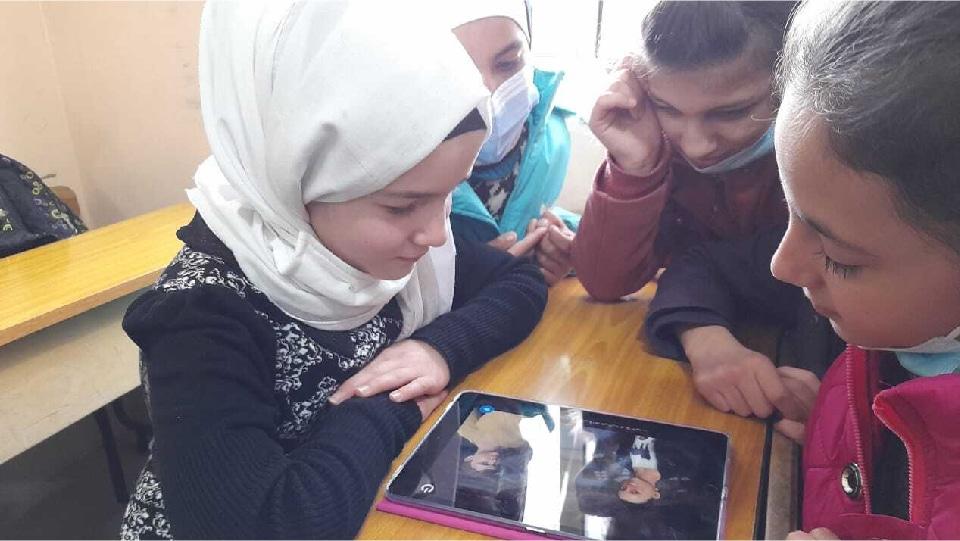 Mädchen mit Kopftüchern sitzen im Klassenzimmer um ein Tablet und spielen Helping Hand