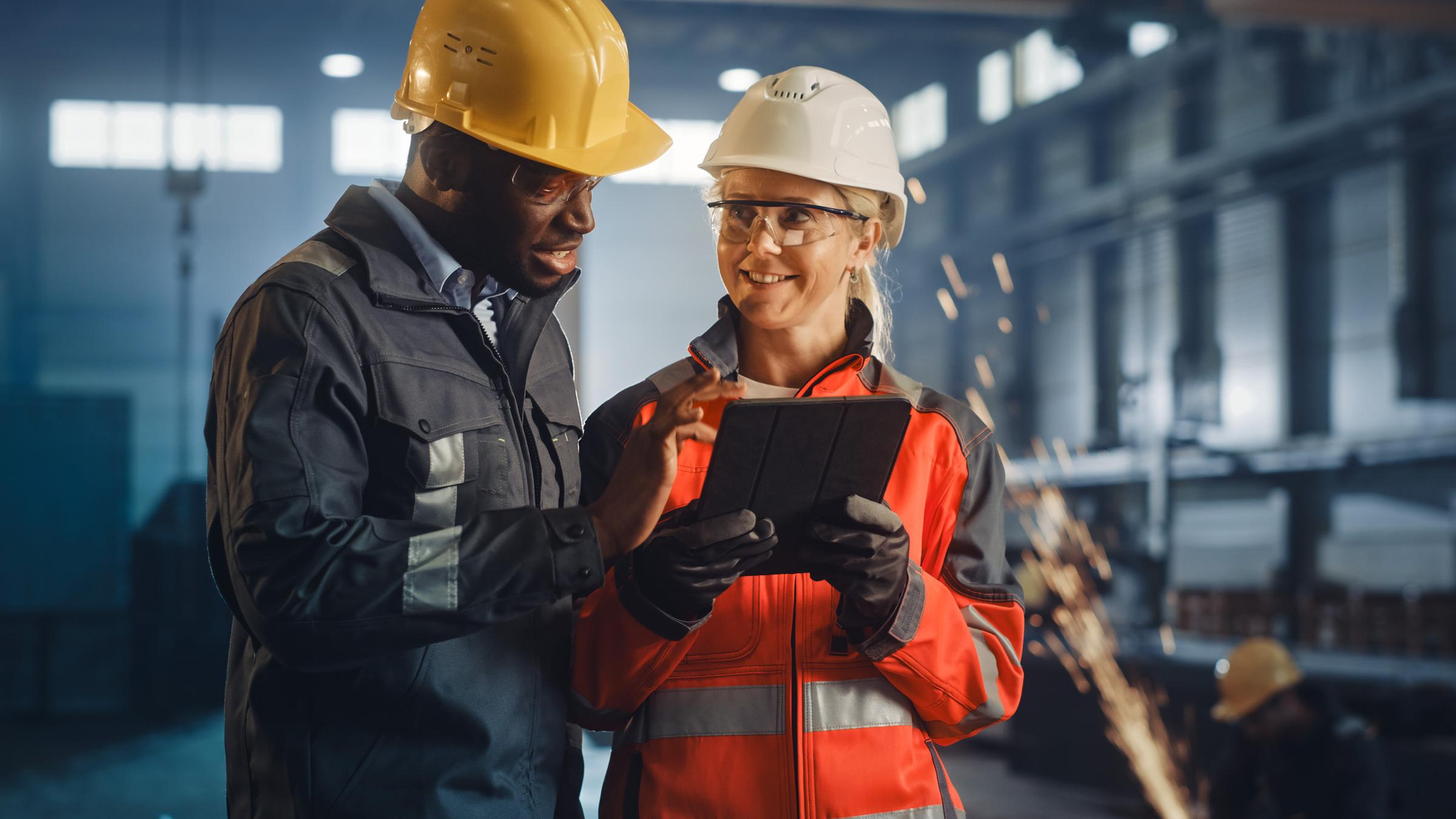 Zwei Arbeitende mit Schutzhelmen prüfen ein Tablet in einer Fabrikhalle