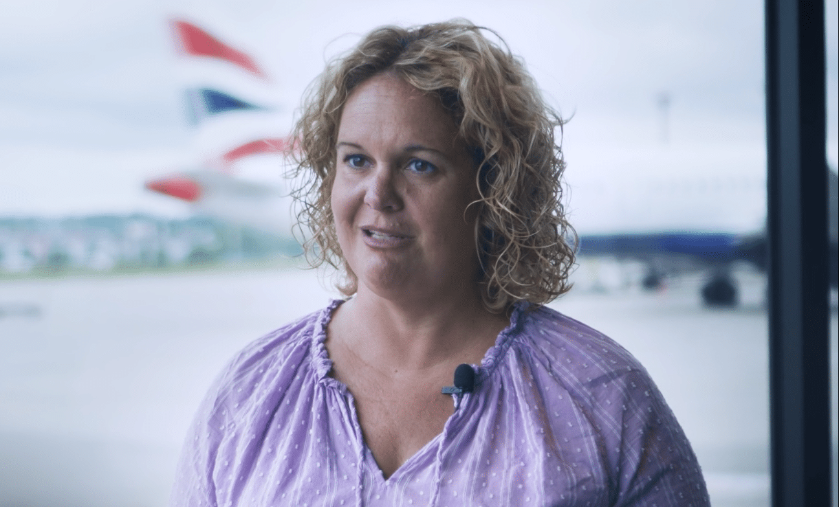 Steph Rosenik von SSP spricht in einem Flughafenterminal in die Kamera, hinter ihr ein Flugzeug