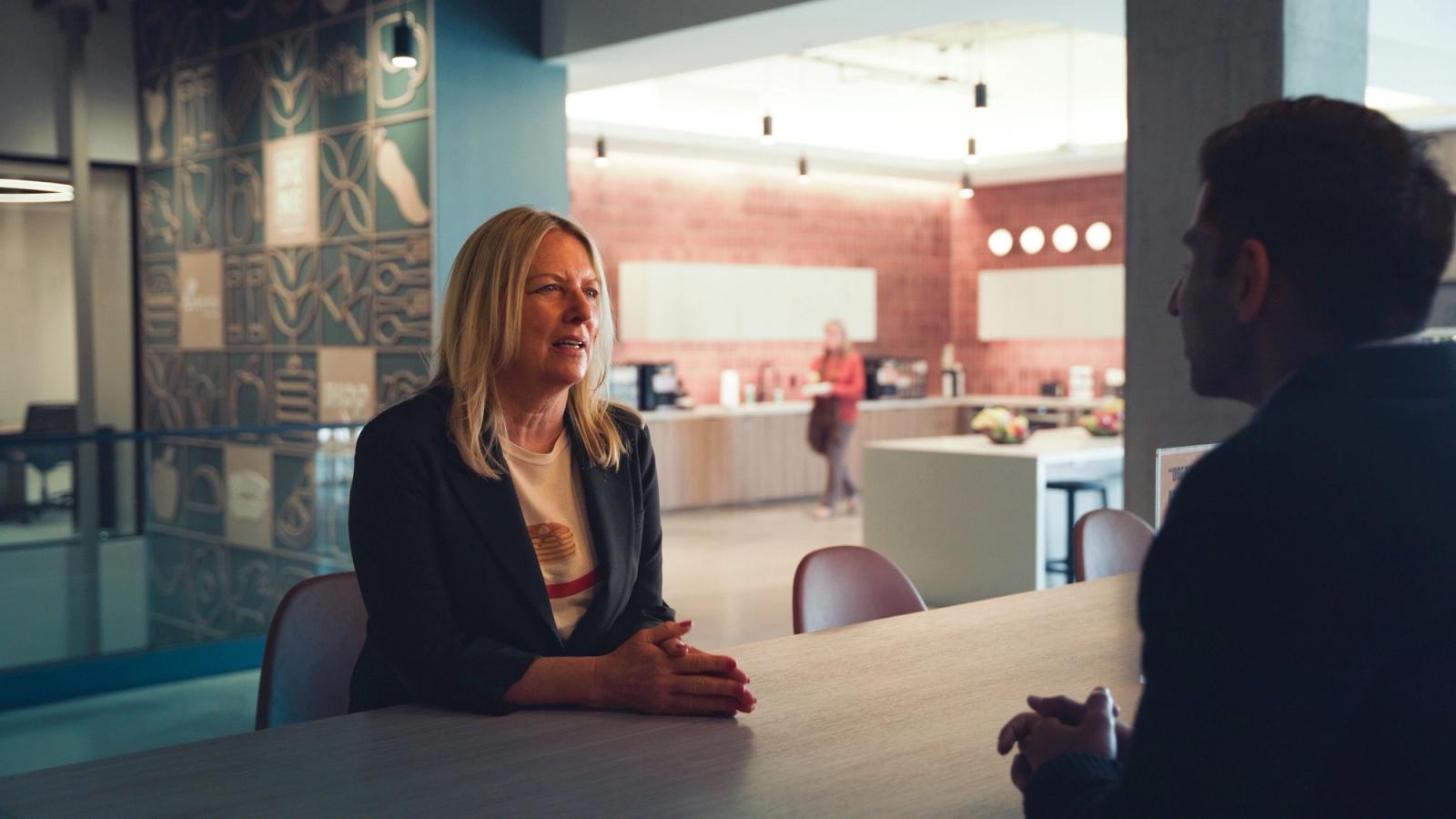 Eine Frau sitzt an einem langen Tisch im Gespräch mit einem Mann, der von hinten zu sehen ist, in einem Café-Bereich
