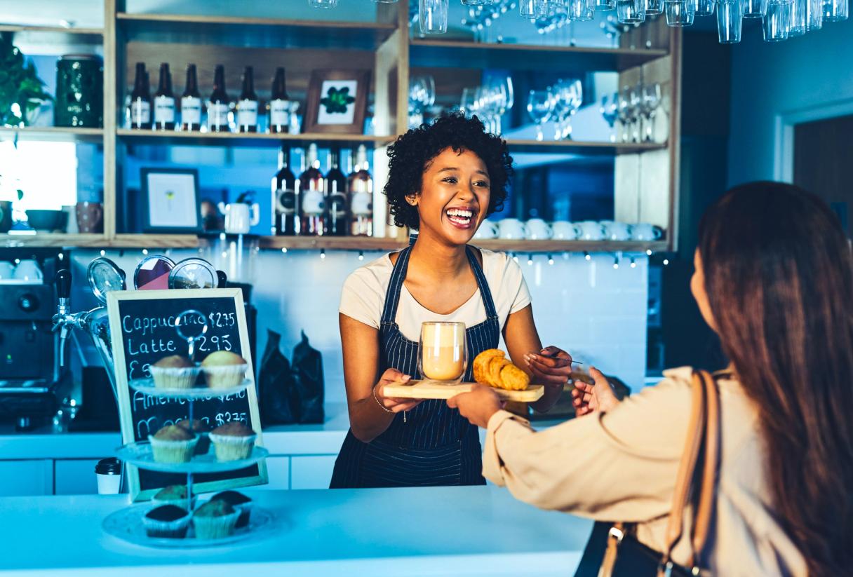 Eine lächelnde Barista reicht einem Gast an der Café-Theke einen Latte und ein Croissant.